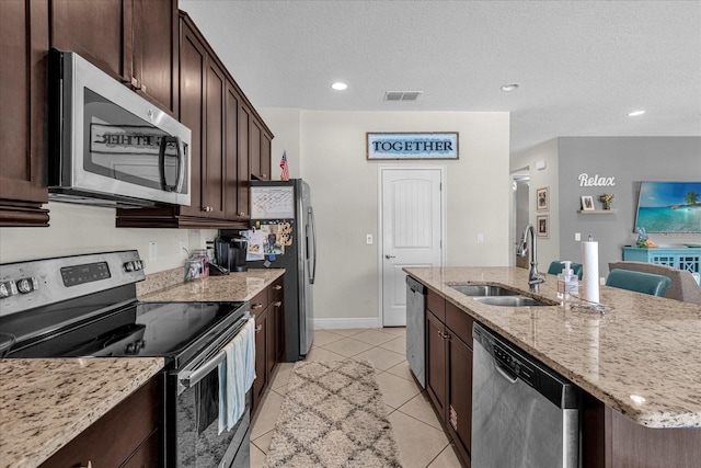 kitchen featuring appliances with stainless steel finishes, light tile floors, light stone counters, sink, and an island with sink