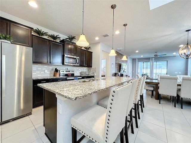 kitchen with light tile floors, backsplash, decorative light fixtures, stainless steel appliances, and a kitchen island with sink