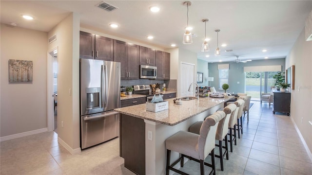 kitchen with pendant lighting, a breakfast bar area, a kitchen island with sink, stainless steel appliances, and light stone countertops