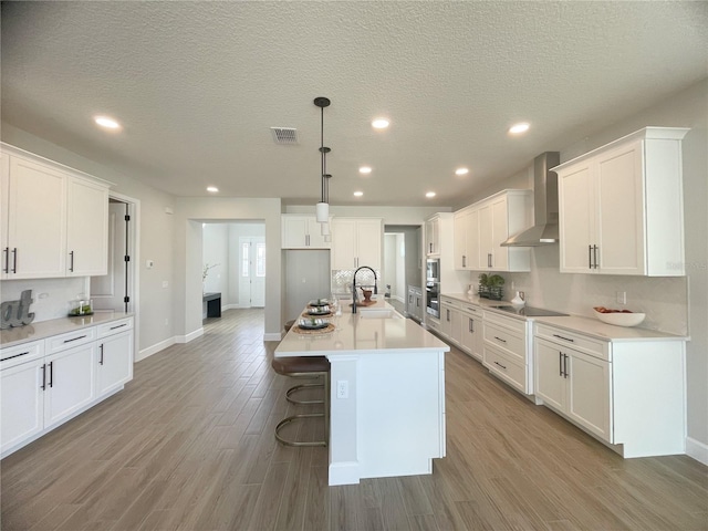 kitchen with white cabinets, hanging light fixtures, light hardwood / wood-style floors, a center island with sink, and wall chimney exhaust hood