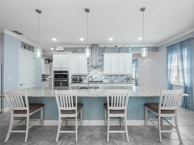 kitchen with crown molding, pendant lighting, wall chimney range hood, white cabinets, and a kitchen island with sink