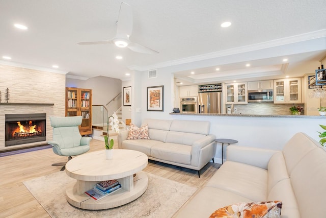 living room with crown molding, ceiling fan, light hardwood / wood-style floors, a textured ceiling, and a stone fireplace