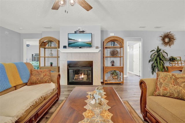 living room with a textured ceiling, wood-type flooring, ceiling fan, and a tiled fireplace