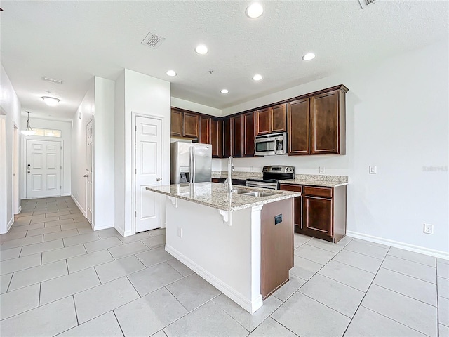 kitchen featuring appliances with stainless steel finishes, dark brown cabinetry, light stone countertops, an island with sink, and light tile patterned floors