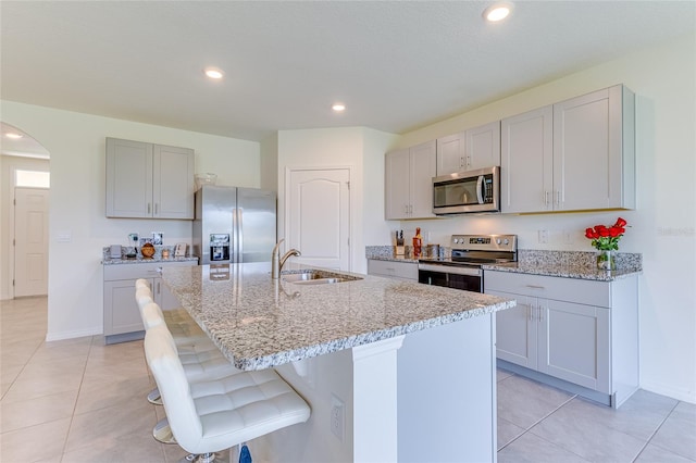 kitchen with sink, a kitchen island with sink, stainless steel appliances, and light stone counters