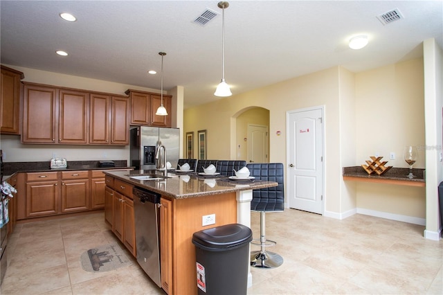 kitchen featuring stainless steel appliances, light tile flooring, a kitchen island with sink, dark stone countertops, and sink