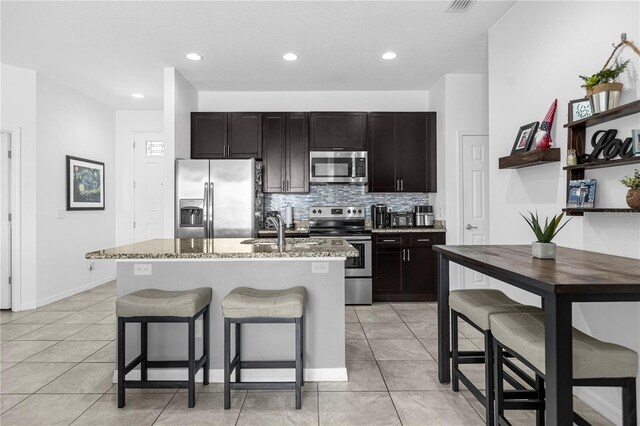 kitchen featuring light stone counters, decorative backsplash, a center island with sink, light tile patterned flooring, and appliances with stainless steel finishes