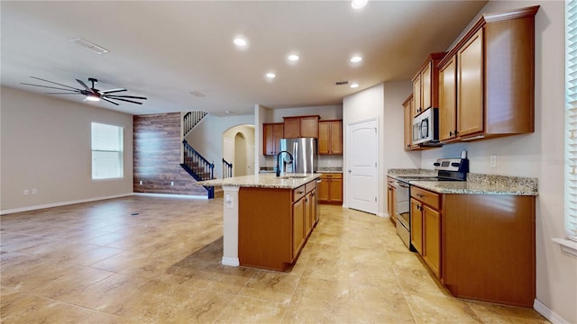 kitchen featuring ceiling fan, sink, light stone countertops, appliances with stainless steel finishes, and an island with sink