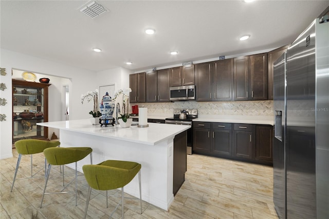 kitchen with stainless steel appliances, a kitchen breakfast bar, a center island with sink, tasteful backsplash, and dark brown cabinets