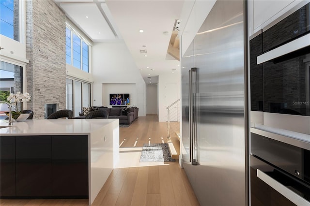 kitchen with appliances with stainless steel finishes, light wood-type flooring, a towering ceiling, white cabinetry, and a stone fireplace