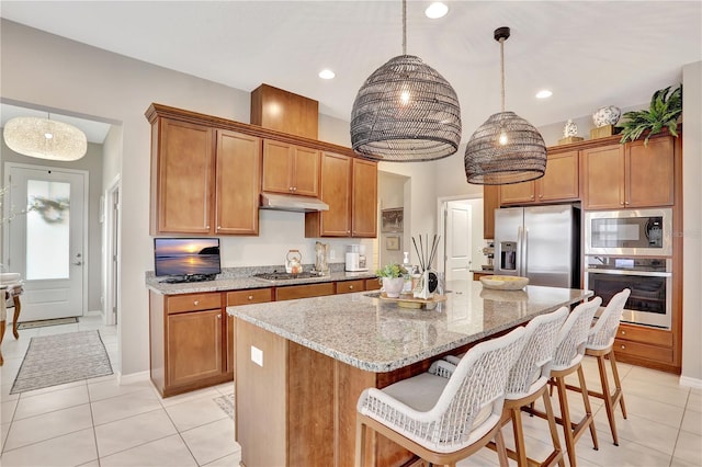 kitchen featuring hanging light fixtures, stainless steel appliances, an island with sink, light stone counters, and light tile floors