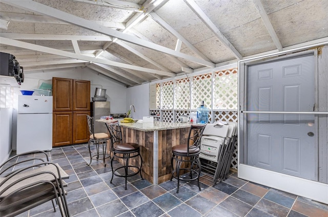 kitchen with a kitchen breakfast bar, white fridge, vaulted ceiling, light stone counters, and dark tile flooring