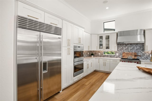 kitchen with white cabinets, crown molding, wall chimney exhaust hood, appliances with stainless steel finishes, and light stone counters