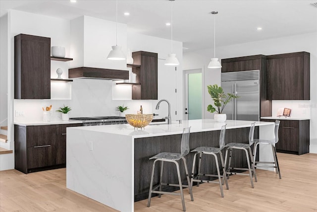 kitchen featuring stainless steel built in refrigerator, dark brown cabinetry, an island with sink, and light hardwood / wood-style floors