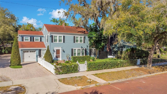 colonial home featuring an attached garage, decorative driveway, a fenced front yard, and a chimney