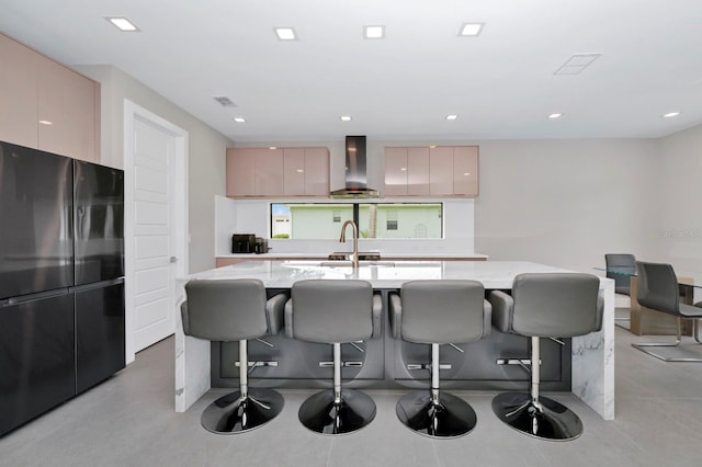 kitchen with a kitchen breakfast bar, black refrigerator, light stone counters, wall chimney range hood, and a kitchen island with sink