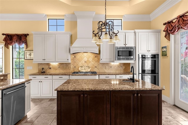 kitchen with appliances with stainless steel finishes, premium range hood, a kitchen island with sink, and a notable chandelier