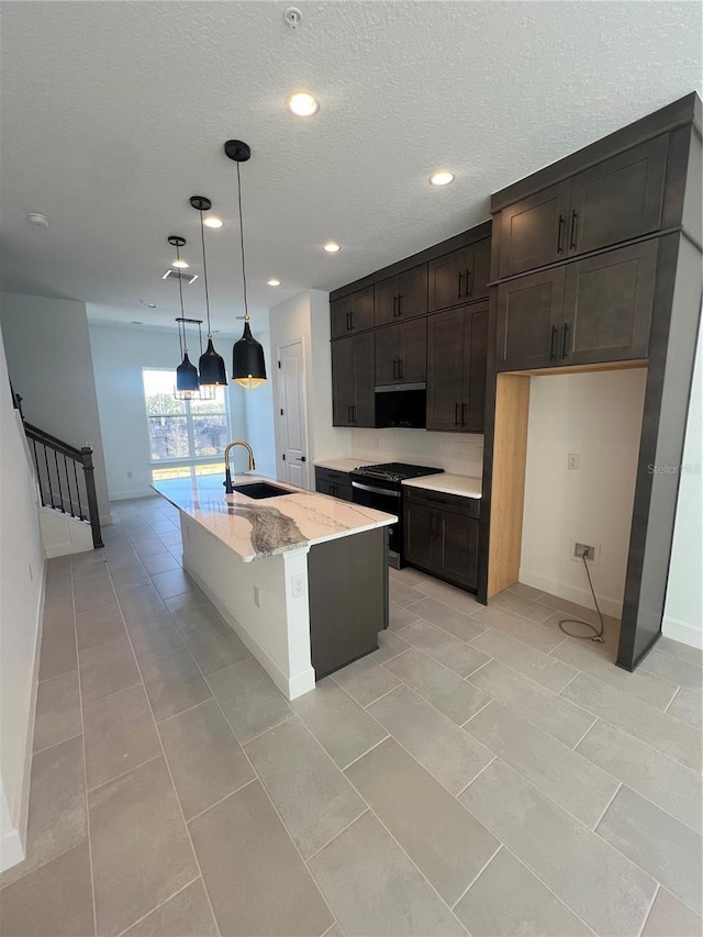 kitchen with sink, hanging light fixtures, a kitchen island with sink, light stone countertops, and gas range oven