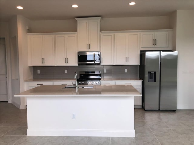 kitchen with an island with sink, backsplash, light tile floors, white cabinets, and appliances with stainless steel finishes