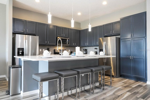 kitchen featuring light wood-type flooring, stainless steel appliances, an island with sink, decorative light fixtures, and backsplash