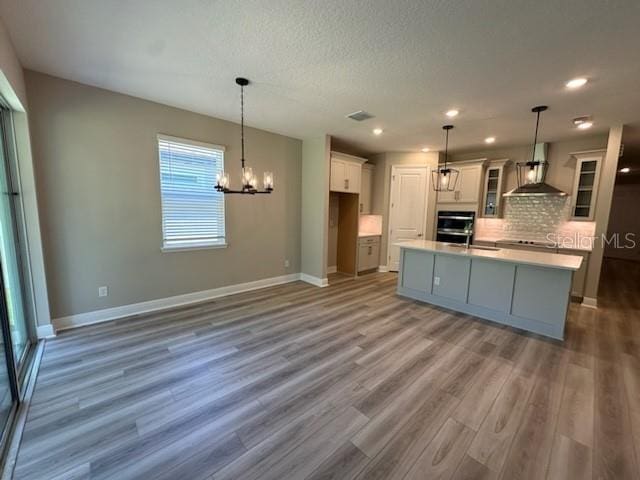 kitchen with decorative light fixtures, wood-type flooring, backsplash, wall chimney exhaust hood, and a kitchen island with sink