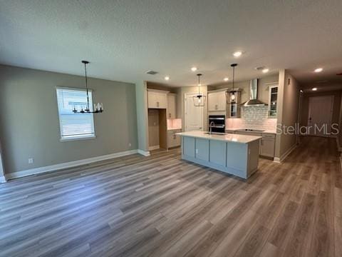 kitchen with hanging light fixtures, a center island with sink, hardwood / wood-style flooring, tasteful backsplash, and wall chimney exhaust hood