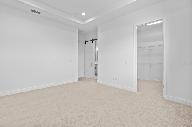 unfurnished bedroom featuring a closet, a tray ceiling, light carpet, a barn door, and a walk in closet