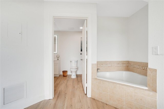 bathroom with a relaxing tiled tub, hardwood / wood-style flooring, toilet, and electric panel