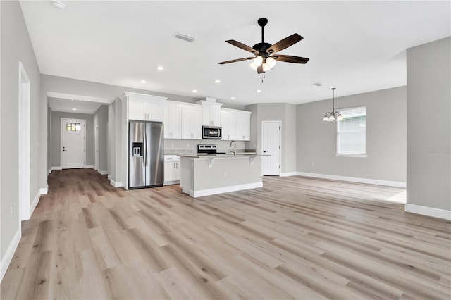 kitchen with sink, ceiling fan with notable chandelier, stainless steel appliances, white cabinets, and an island with sink