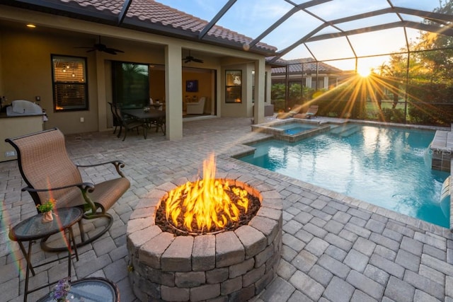 pool at dusk featuring glass enclosure, a fire pit, an in ground hot tub, a patio area, and ceiling fan