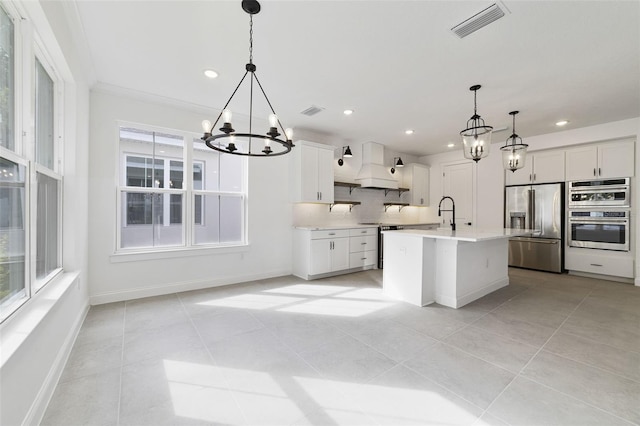 kitchen with pendant lighting, premium range hood, a kitchen island with sink, stainless steel appliances, and white cabinets