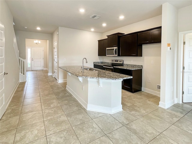 kitchen featuring a center island with sink, sink, appliances with stainless steel finishes, dark brown cabinets, and light stone counters