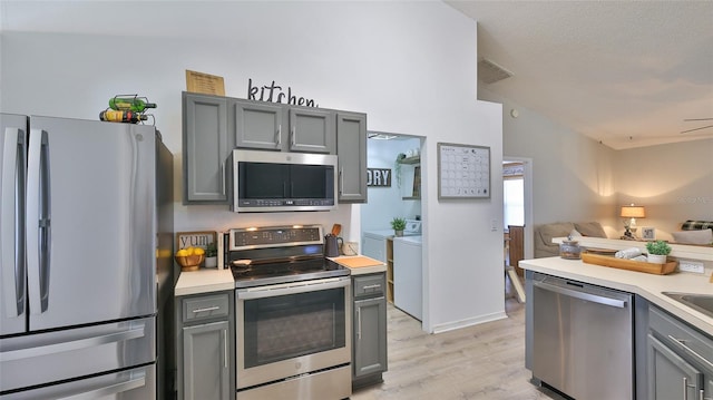 kitchen with appliances with stainless steel finishes, gray cabinets, independent washer and dryer, ceiling fan, and light hardwood / wood-style flooring