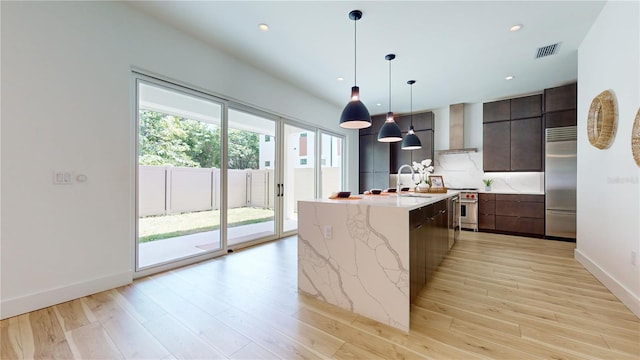 kitchen with light hardwood / wood-style flooring, pendant lighting, wall chimney exhaust hood, high end appliances, and a kitchen island with sink