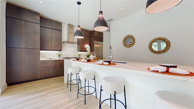 kitchen with light hardwood / wood-style floors, sink, a kitchen breakfast bar, wall chimney exhaust hood, and decorative light fixtures