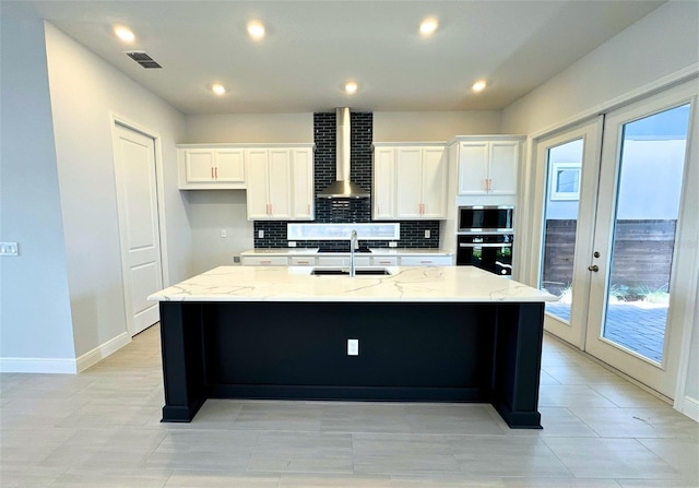 kitchen featuring wall chimney range hood, french doors, white cabinets, and a center island with sink