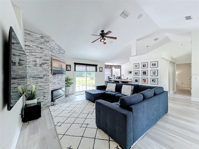 living room with ceiling fan, a stone fireplace, light hardwood / wood-style floors, vaulted ceiling, and a textured ceiling