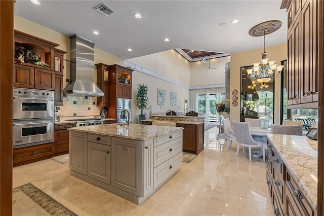 kitchen with a kitchen island with sink, an inviting chandelier, wall chimney exhaust hood, a textured ceiling, and stainless steel appliances