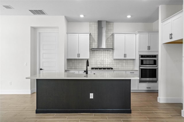 kitchen featuring a center island with sink, white cabinetry, stainless steel appliances, and wall chimney range hood