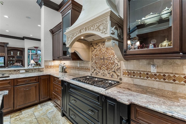 kitchen featuring backsplash, dark brown cabinets, ornamental molding, light stone countertops, and stainless steel gas cooktop