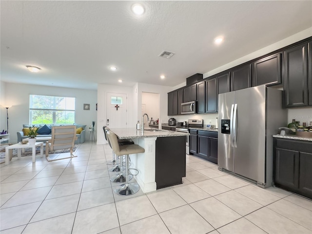 kitchen with sink, light stone countertops, a center island with sink, a breakfast bar, and appliances with stainless steel finishes