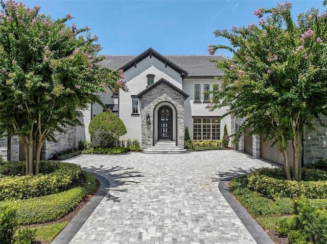 french provincial home with a tile roof, stucco siding, decorative driveway, a garage, and stone siding