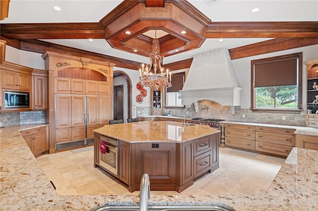 kitchen featuring light stone countertops, premium range hood, black microwave, decorative light fixtures, and a center island with sink