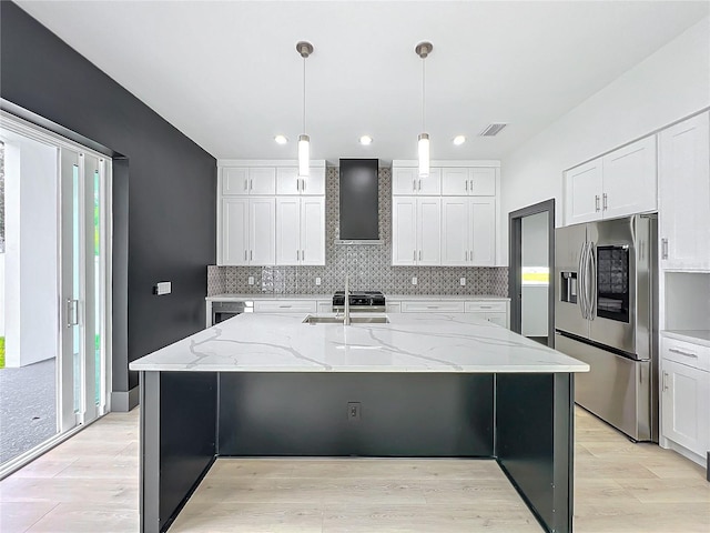 kitchen featuring wall chimney exhaust hood, stainless steel fridge, an island with sink, and hanging light fixtures