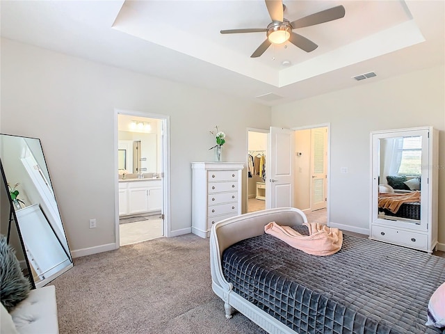 carpeted bedroom with connected bathroom, a tray ceiling, a walk in closet, and ceiling fan