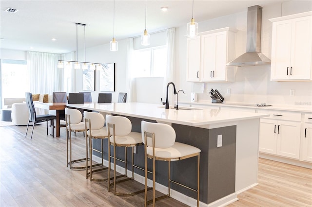 kitchen featuring a center island with sink, white cabinets, a healthy amount of sunlight, and wall chimney range hood