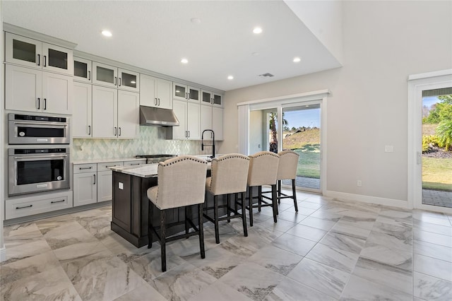 kitchen featuring light stone countertops, decorative backsplash, stainless steel double oven, a breakfast bar area, and an island with sink