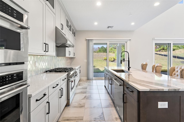 kitchen with a kitchen island with sink, sink, light stone countertops, white cabinetry, and stainless steel appliances