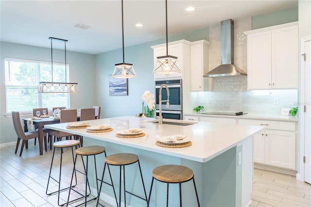 kitchen with pendant lighting, stainless steel double oven, a center island with sink, and wall chimney range hood