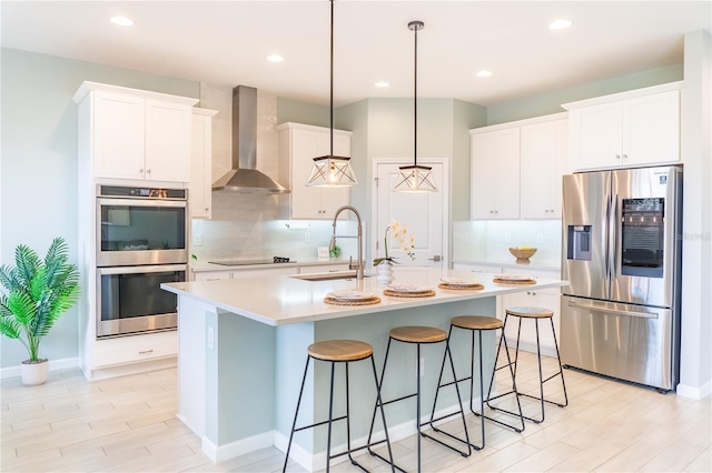 kitchen with hanging light fixtures, white cabinetry, stainless steel appliances, a center island with sink, and wall chimney range hood